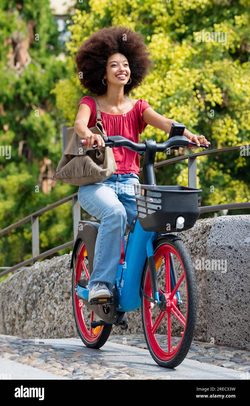 Una donna nera soddisfatta con i suoi acconciature afro in bicicletta e con lo sguardo lontano sulla strada urbana di giorno Foto Stock