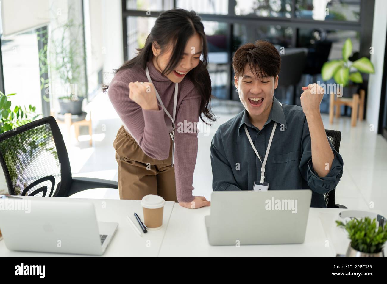Due giovani e felici impiegati asiatici guardano lo schermo di un notebook, rallegrandosi e celebrando il successo del loro progetto in ufficio Foto Stock