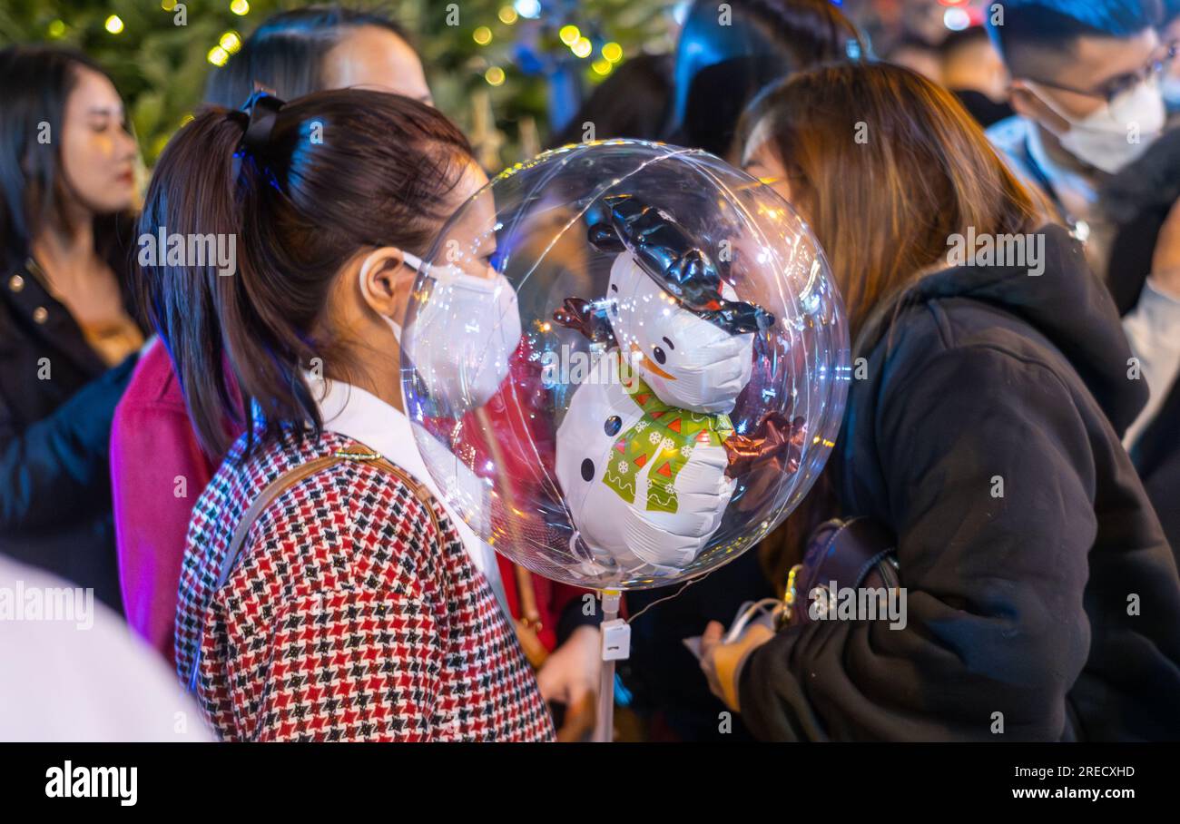 Una giovane donna vietnamita porta con sé una mongolfiera chiara con un pupazzo di neve bianco all'interno in fitta folla di notte durante l'inverno vicino al Natale ad Hanoi, Vietna Foto Stock