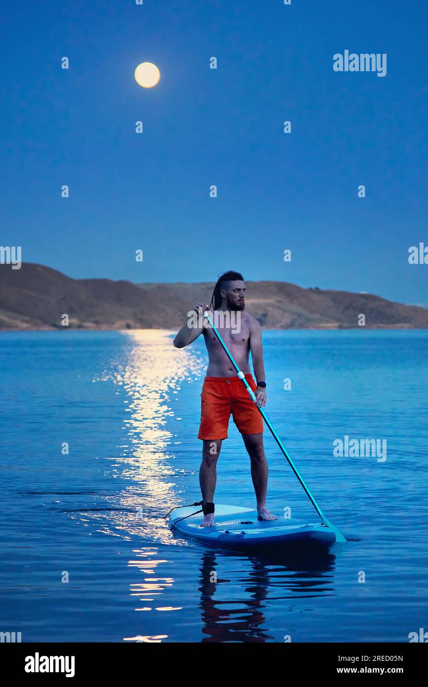 Uomo surfista in camicia arancione alle tavole SUP in piedi con silhouette contro il percorso lunare di luna piena vicino all'isola del lago Kapchagay in Kazakistan. Foto Stock