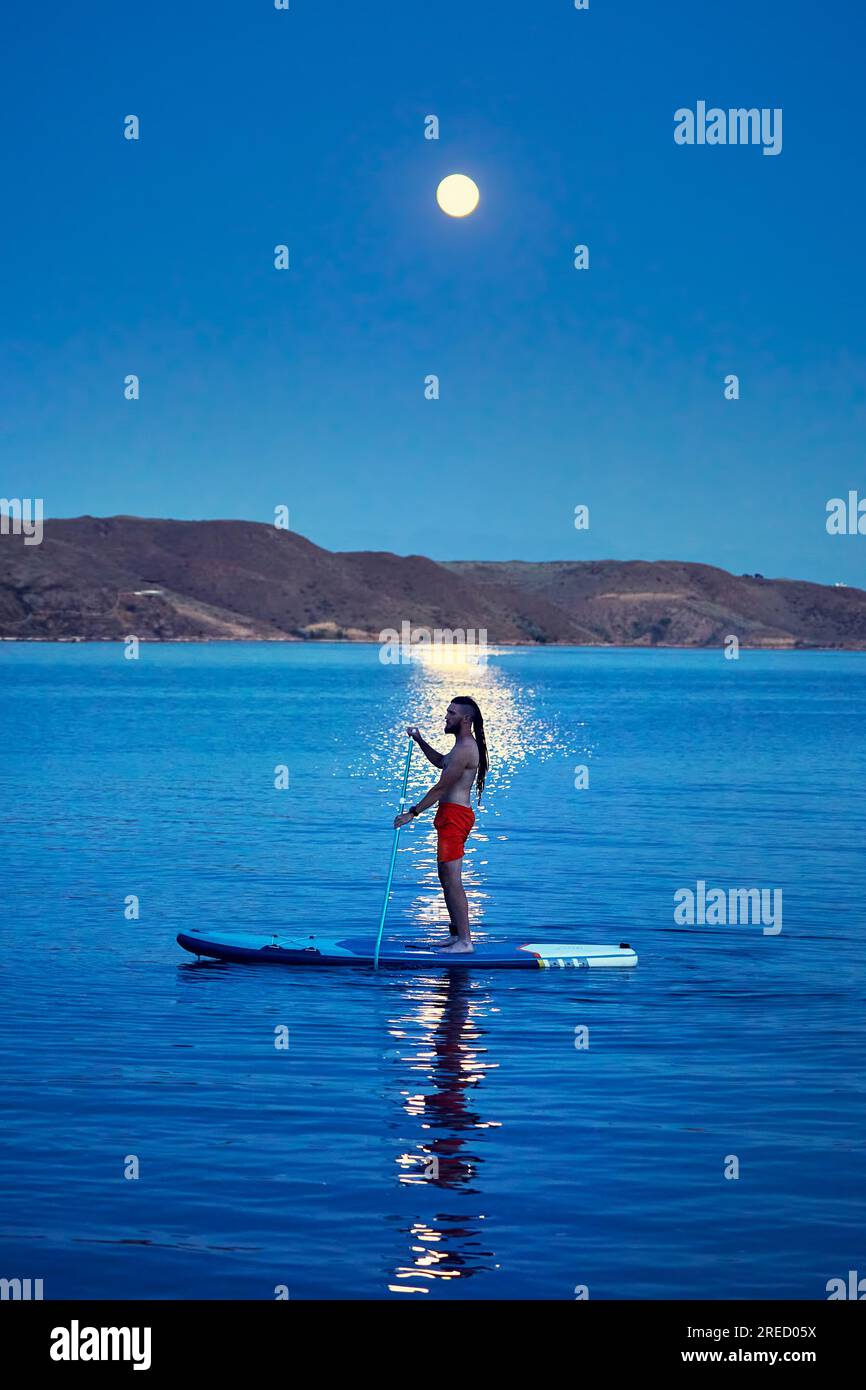 Uomo surfista in camicia arancione alle tavole SUP in piedi con silhouette contro il percorso lunare di luna piena vicino all'isola del lago Kapchagay in Kazakistan. Foto Stock