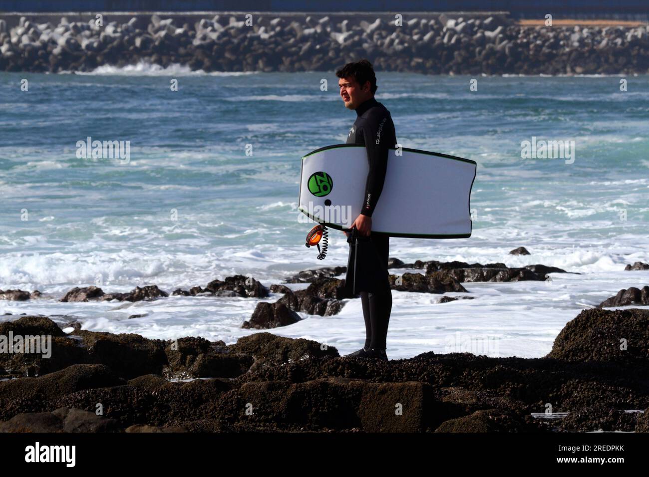 Bodyboarder in piedi su rocce che guardano al mare sull'Oceano Pacifico, Arica, Cile Foto Stock