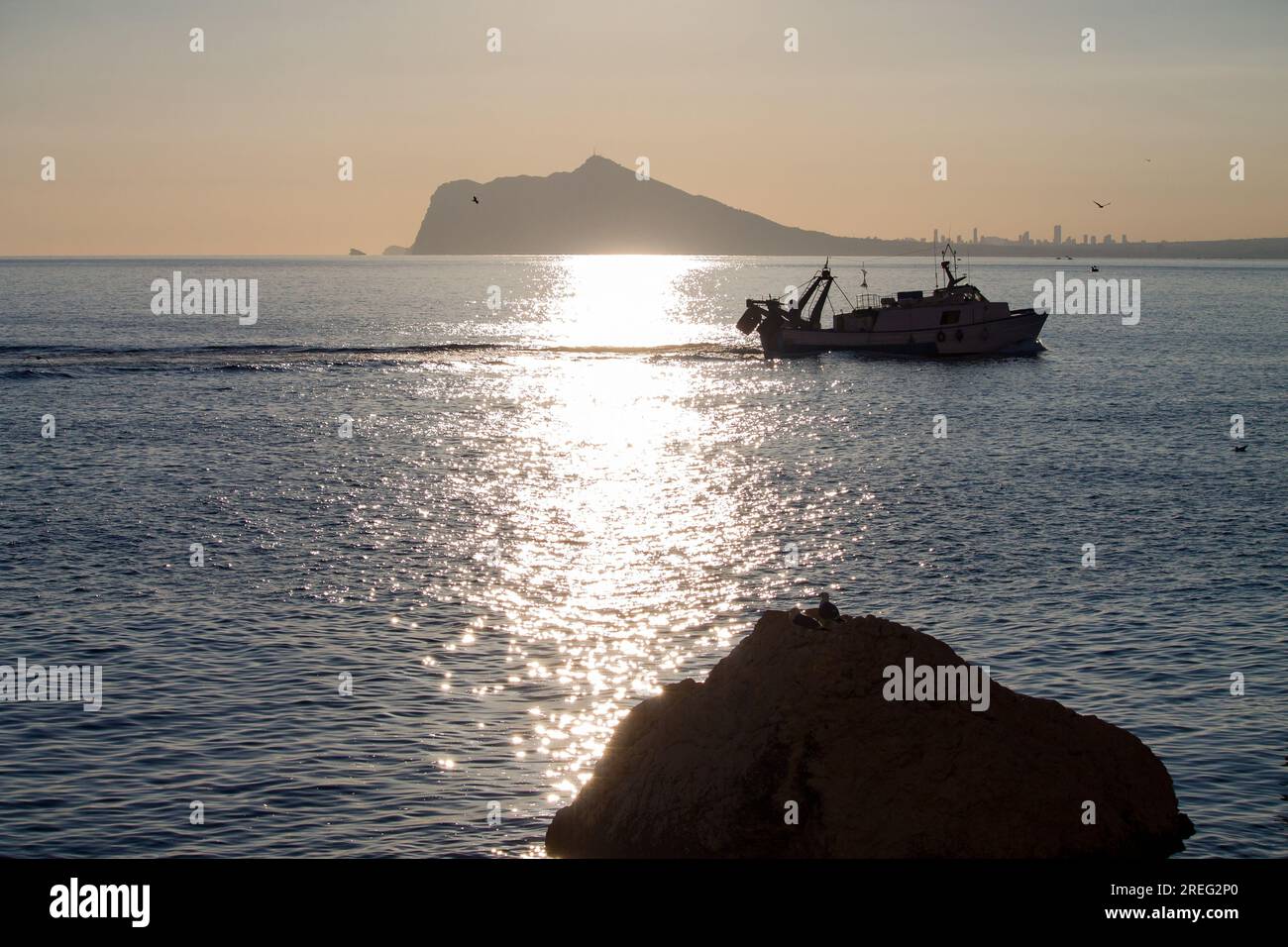Sagome nel Mediterraneo di un peschereccio circondato da gabbiani e strati delle montagne Calpe Foto Stock
