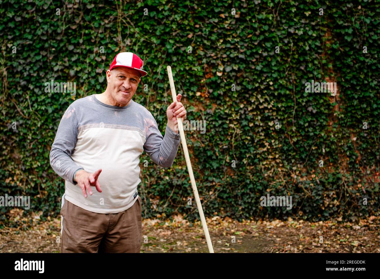 Un anziano sorridente si trova con un rastrello in mano davanti al muro di ivy Foto Stock