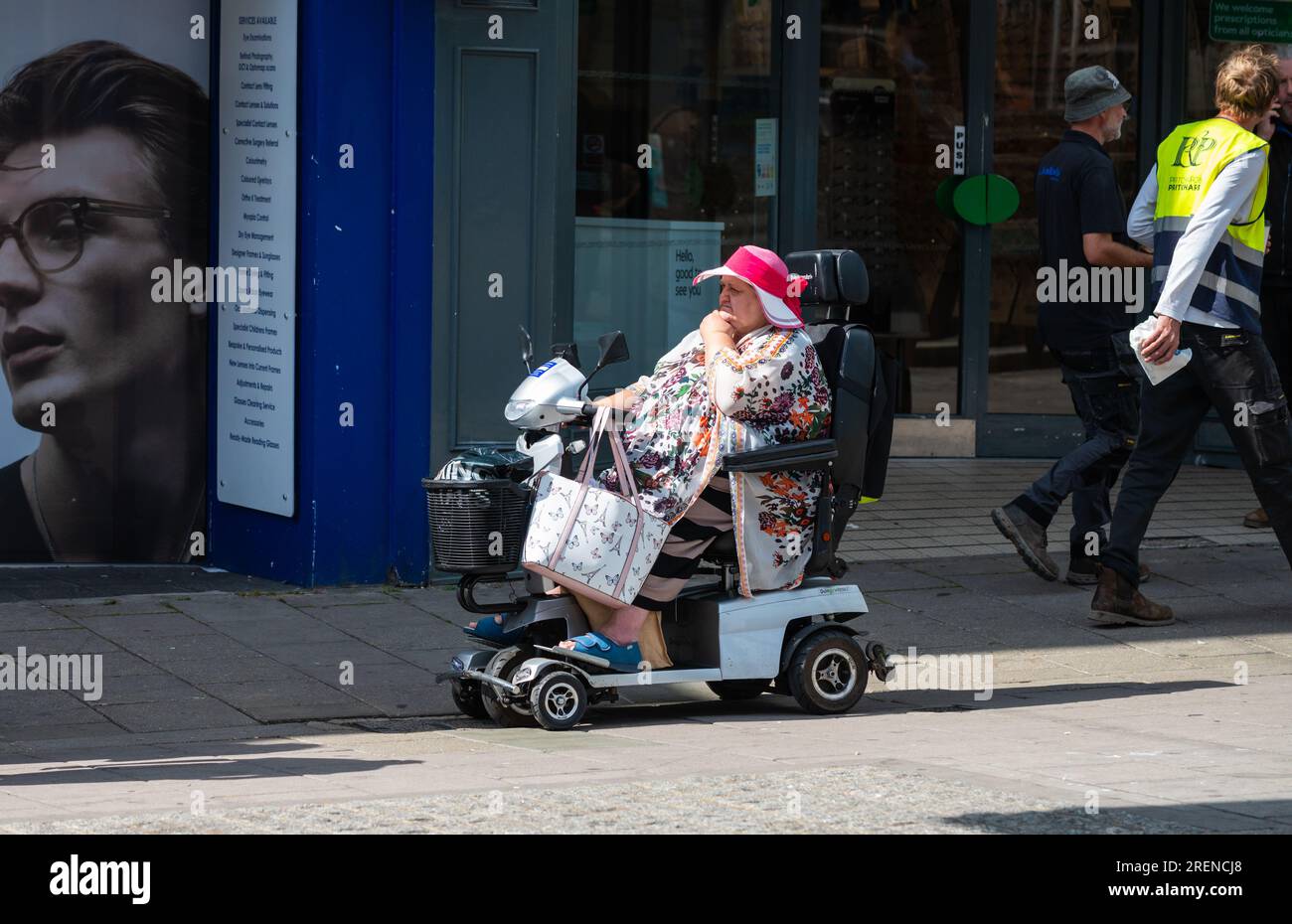 Donna anziana di grandi dimensioni in scooter elettrico per la mobilità sul marciapiede dei negozi, che porta borse per la spesa, indossa abiti estivi e cappello in una città in Inghilterra, Regno Unito. Foto Stock