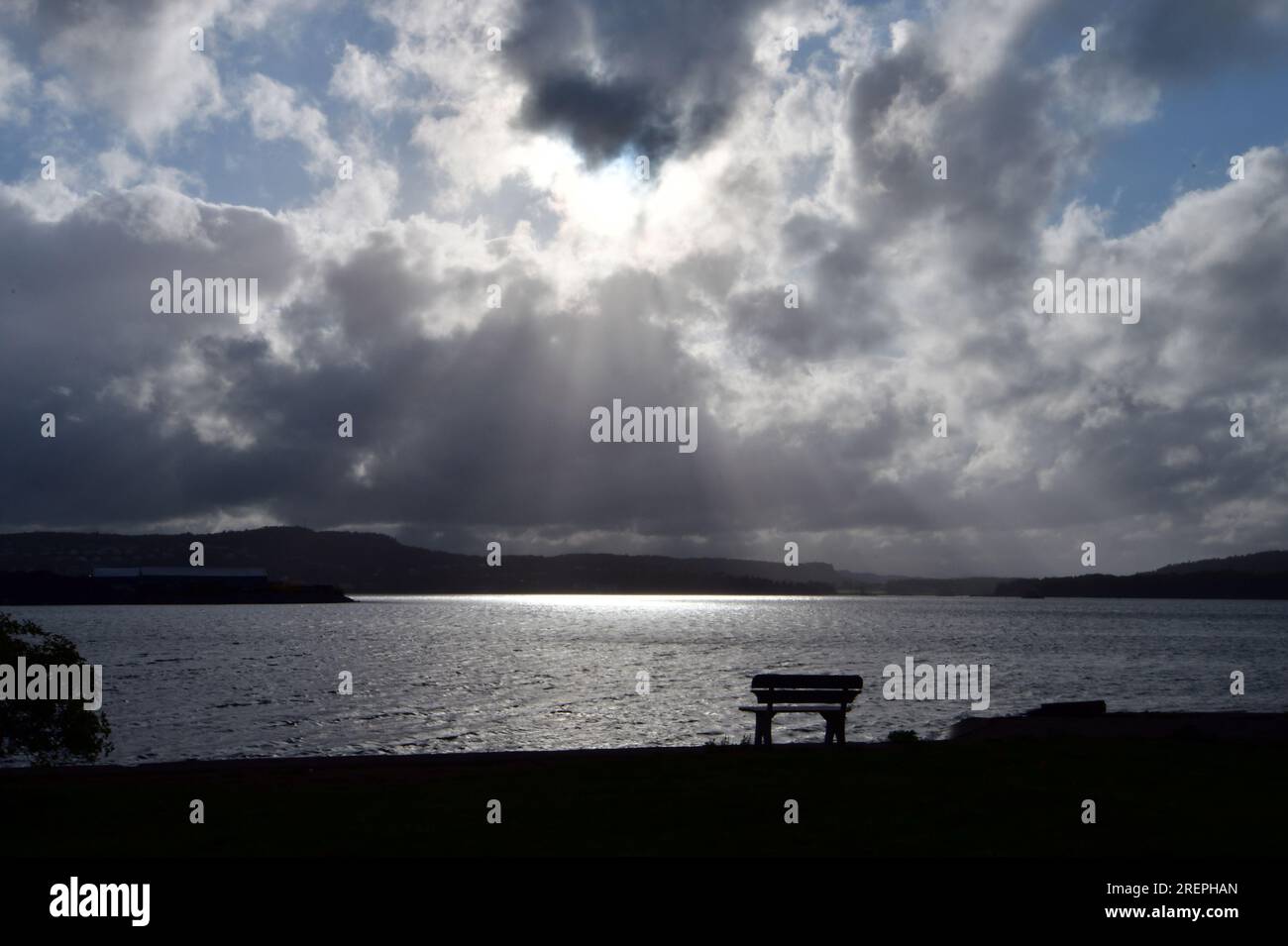 Sagoma di panchina di fronte all'oceano al largo del porto interno di Horten, Norvegia. Raggi del sole attraverso le nuvole contro il cielo azzurro. Foto Stock