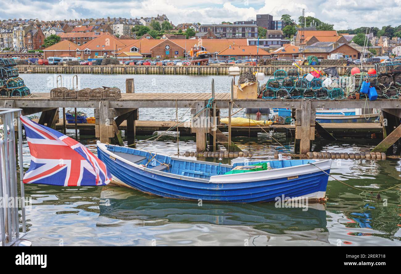 Una barca a remi è ormeggiata su un molo impilato con vasi di aragosta. Gli edifici si trovano sulla riva opposta. Una bandiera dell'unione è in primo piano. Foto Stock