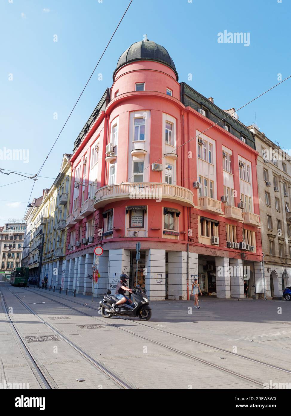 Il motociclista attraversa i binari del tram di fronte a un imponente edificio rosso con cupola all'angolo. Sofia, Bulgaria. 29 luglio 2023 Foto Stock