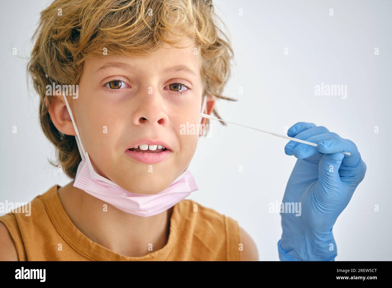 Ritaglia il medico anonimo con il tampone che tocca la guancia di un bambino sensibile che guarda la fotocamera durante il test del coronavirus su sfondo chiaro Foto Stock
