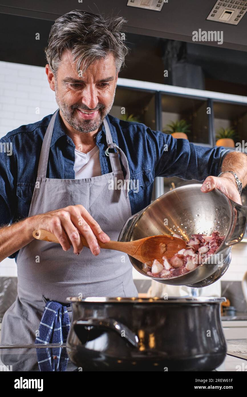 Uno chef maschio positivo in grembiule in piedi al piano cottura e versare il polpo nella pentola mentre si cucina in cucina a casa Foto Stock