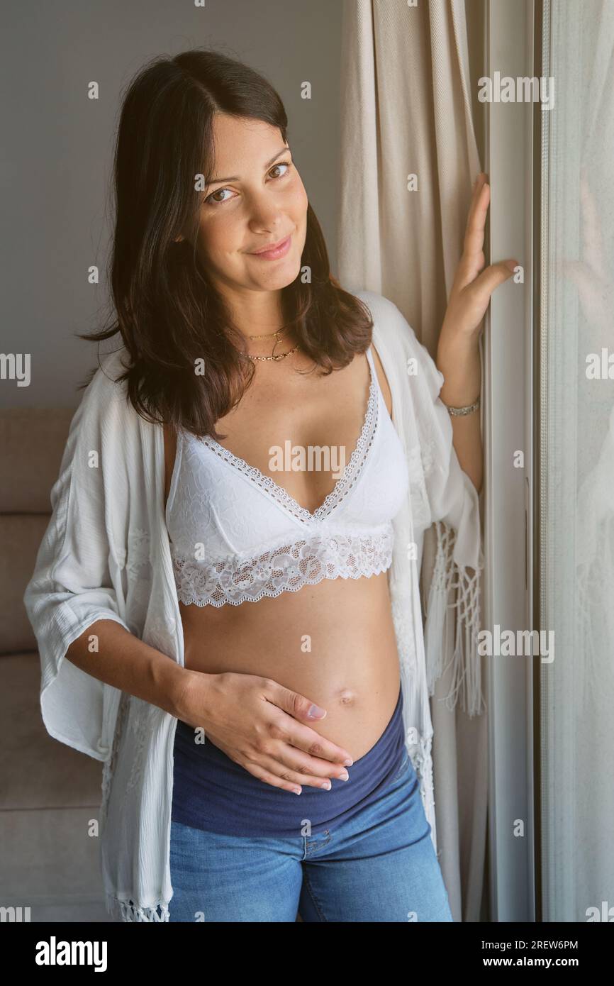 Giovane donna incinta con reggiseno bianco e jeans sorridente e accarezzante di dolcezza mentre si trova vicino alla finestra nel salotto di casa Foto Stock