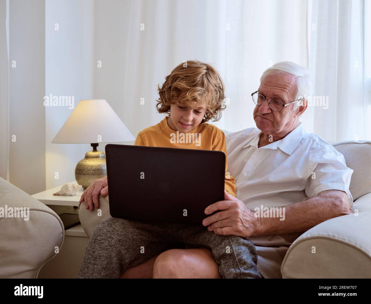 Simpatico nipote che naviga in un netbook moderno mentre si siede seduti su un giro di nonno con gli occhiali da vista in poltrona nel salotto di casa Foto Stock