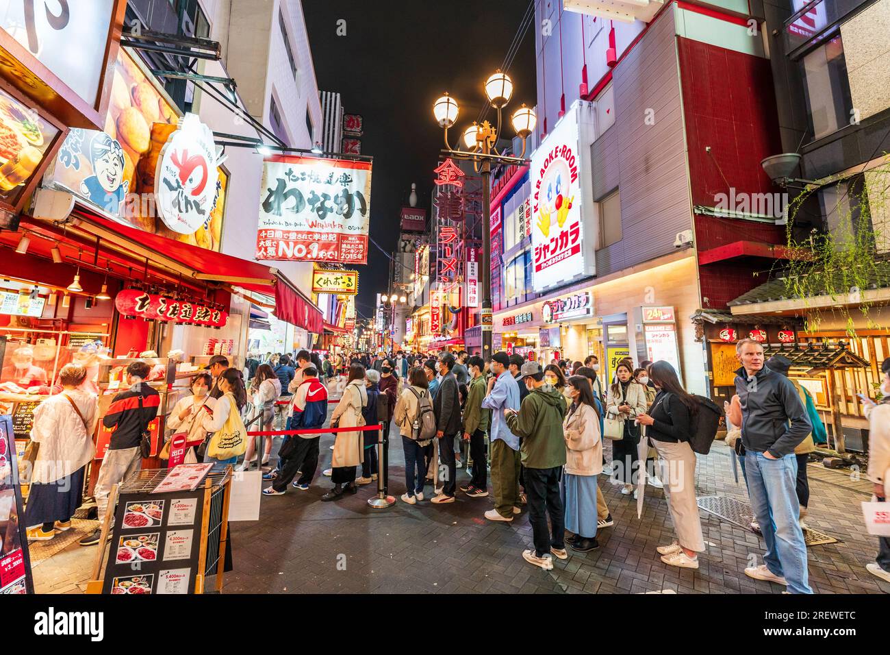 Vista lungo una trafficata e vivace via Dotonbori notturna, Osaka, con la gente in coda al banco take away del ristorante Takoyaki Doraku Wanaka Foto Stock