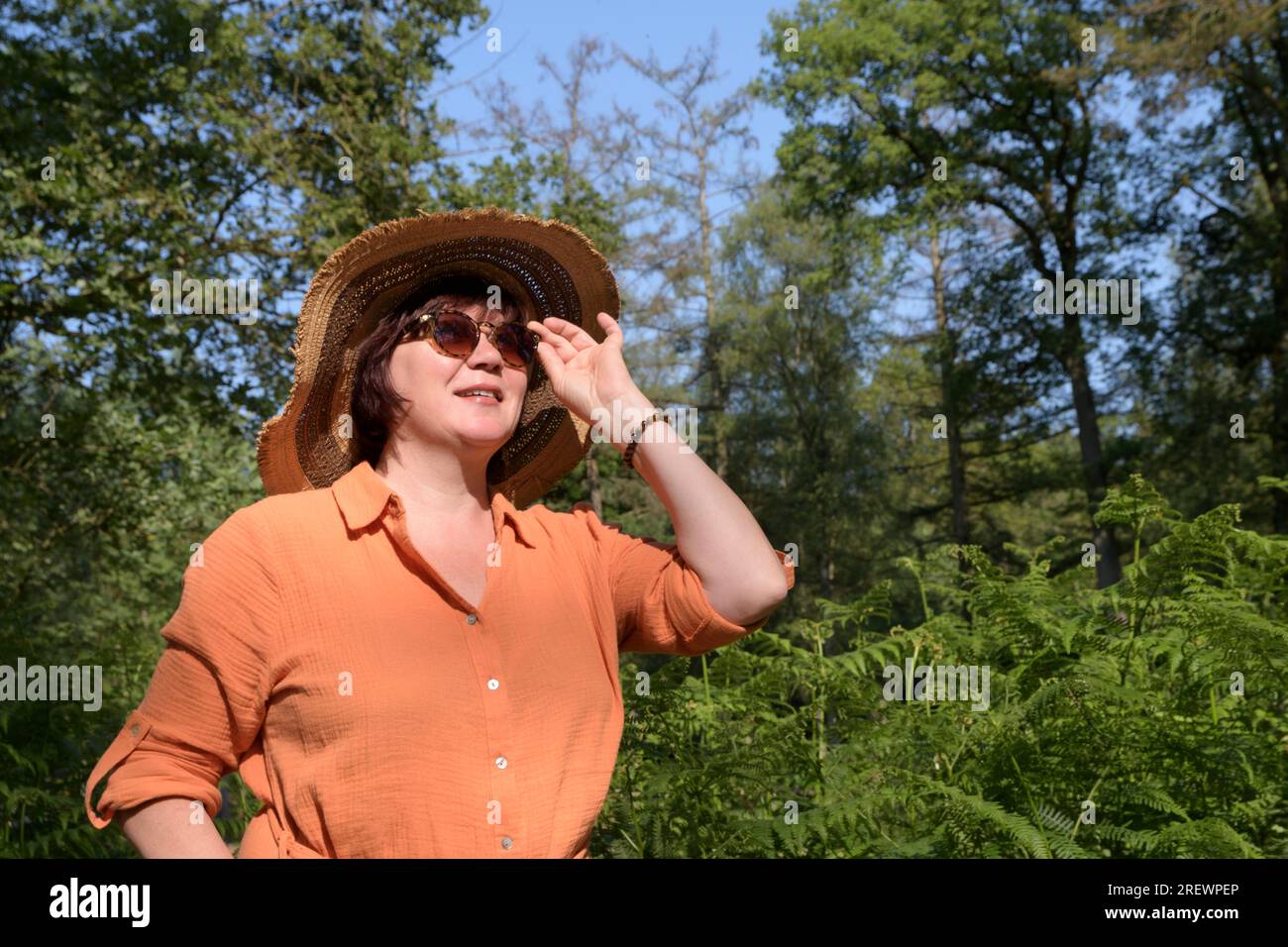 Una donna in un parco ombreggiato. Vestito arancione, cappello di paglia, occhiali da sole. Posiziona per il testo Foto Stock