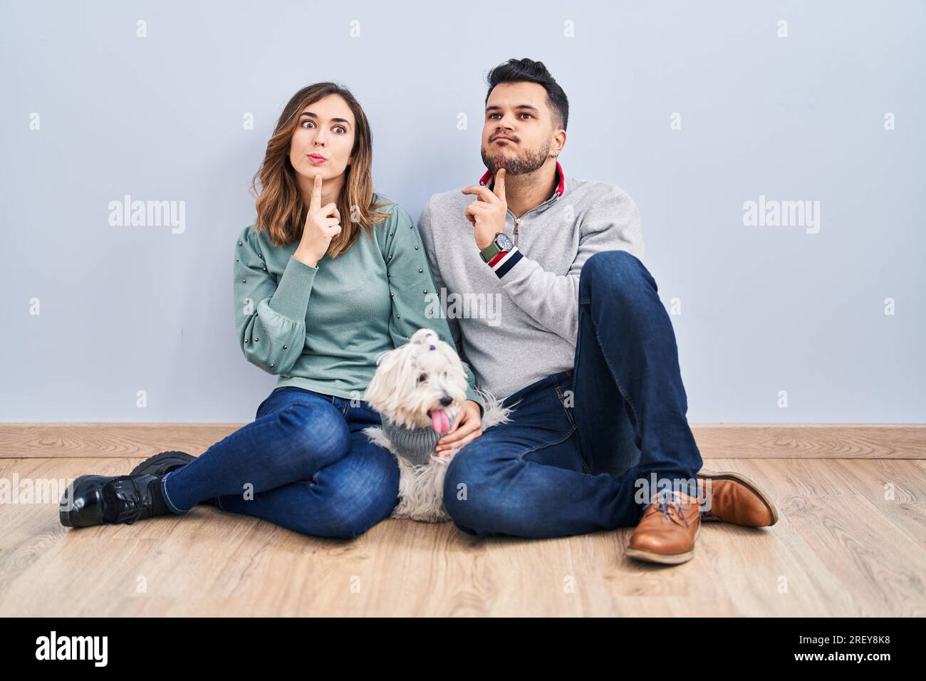 Giovane coppia ispanica seduta sul pavimento con un cane che pensa si concentrò sul dubbio con il dito sul mento e guardando in alto a chiedersi Foto Stock