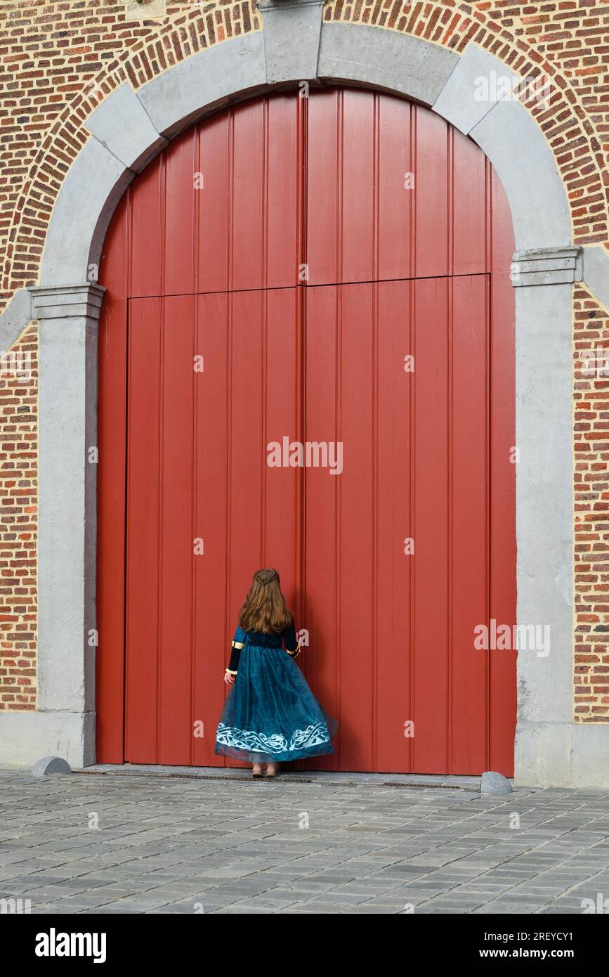 Una bambina in abito medievale bussa alle grandi porte rosse di un antico castello. formato verticale Foto Stock