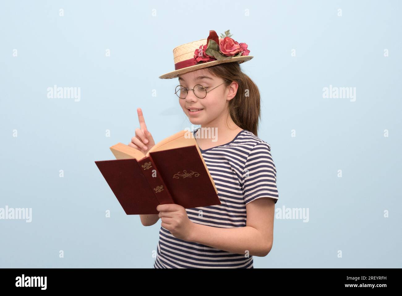 Una ragazza con abiti in stile retrò legge con entusiasmo un libro un cappello di paglia con fiori, occhiali e un libro in mano. Foto Stock