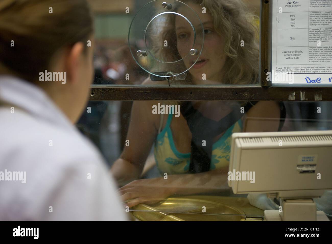 Una donna parla con un impiegato in una biglietteria, separata da una barriera di vetro, in un ambiente italiano. Foto Stock