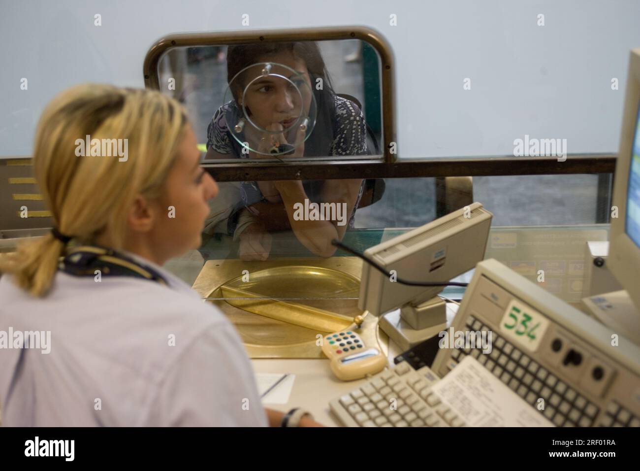 Una donna parla con un'agenzia di biglietteria attraverso un banco di vetro in una stazione o in un aeroporto. Foto Stock