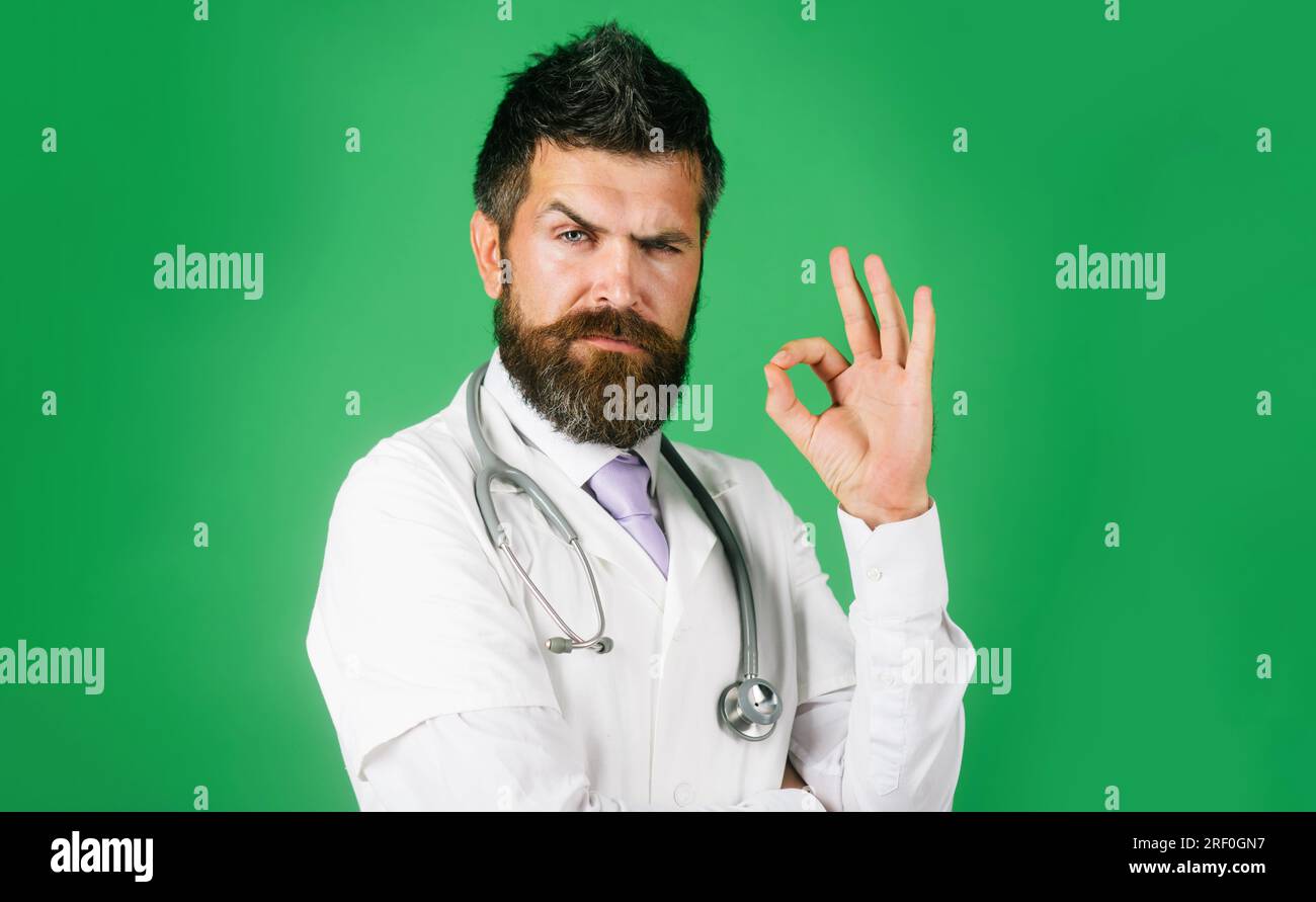 Medico chirurgo che mostra un segno di ok con le dita. Uomo medico barbuto con stetoscopio in camice bianco gestante eccellente. Un serio medico maschio sicuro di sé Foto Stock