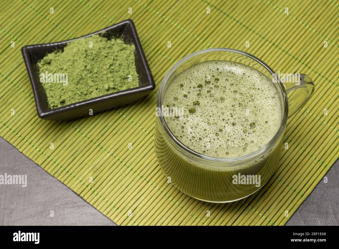 Tè Matcha in un bicchiere, tè matcha in polvere in un recipiente nero su un tovagliolo di bambù verde. Spianatura piatta. Sfondo grigio. Foto Stock