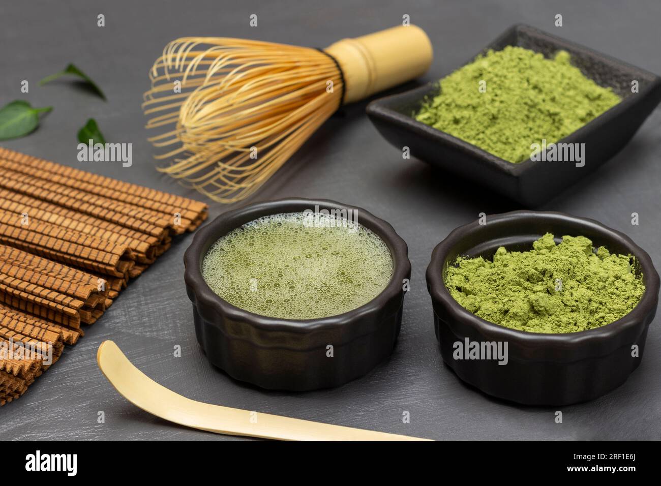 Tè Matcha in polvere e nel tè matcha una ciotola nera. Frusta di bambù e misurino sul tavolo. Sfondo grigio. Foto Stock
