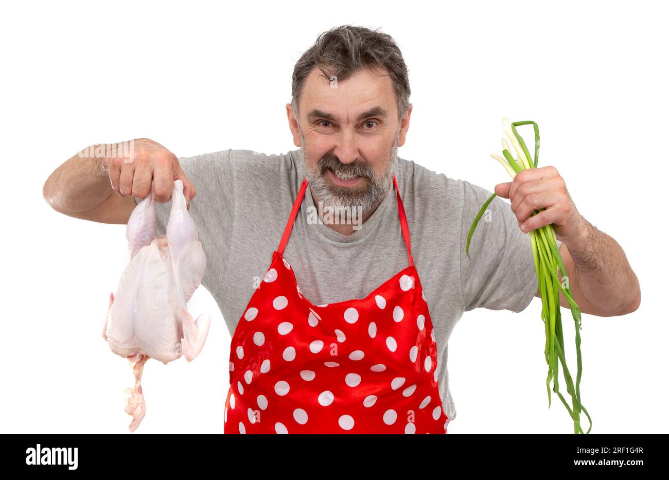 Un anziano cuoco maschio barbuto in grembiule rosso a pois contiene un pollo crudo e un mazzo di cipolle su sfondo bianco. Cuocere con il pollo. Foto Stock