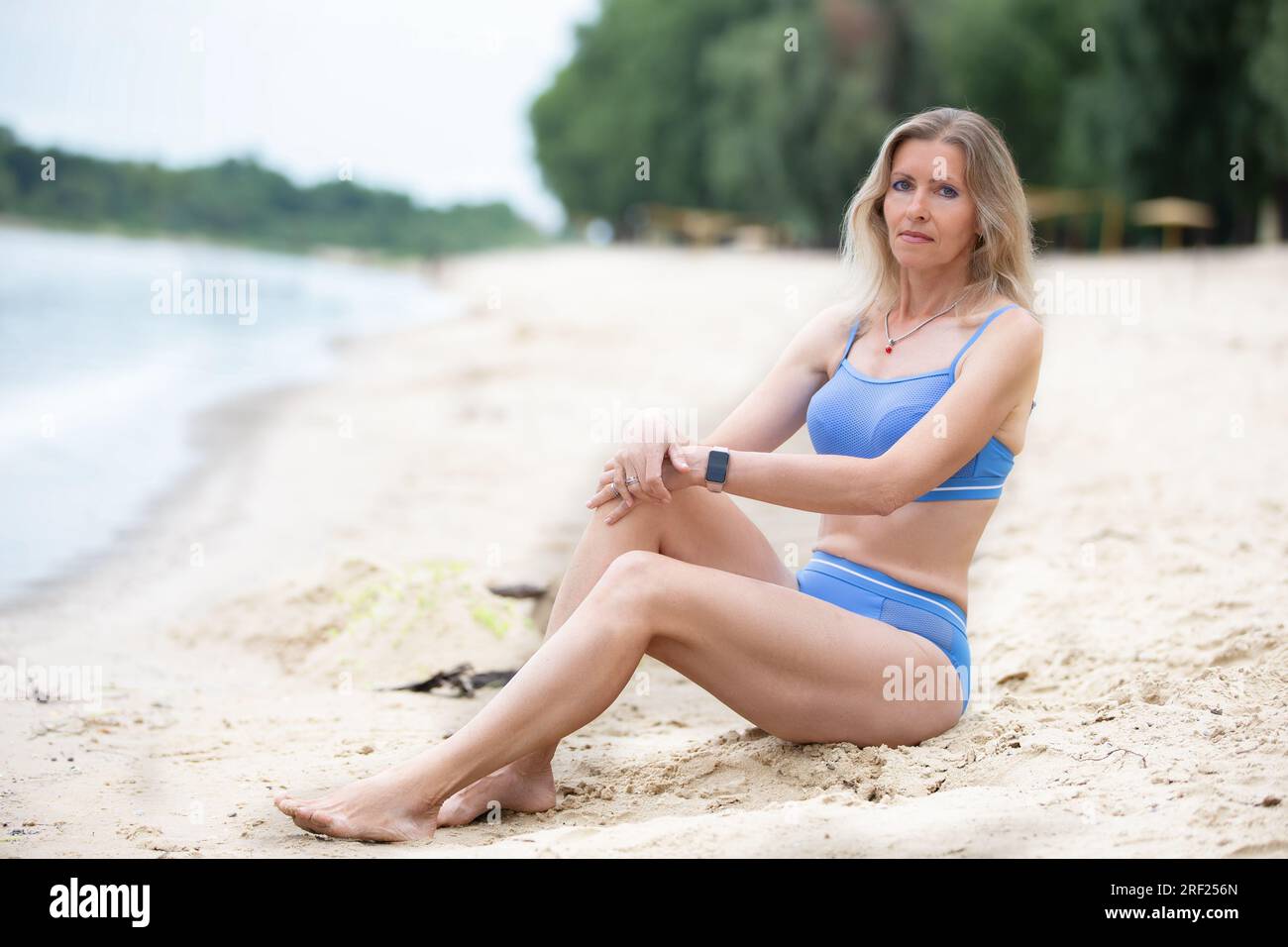 Bella donna bionda magra di mezza età con un costume da bagno si siede sulla spiaggia. Foto Stock