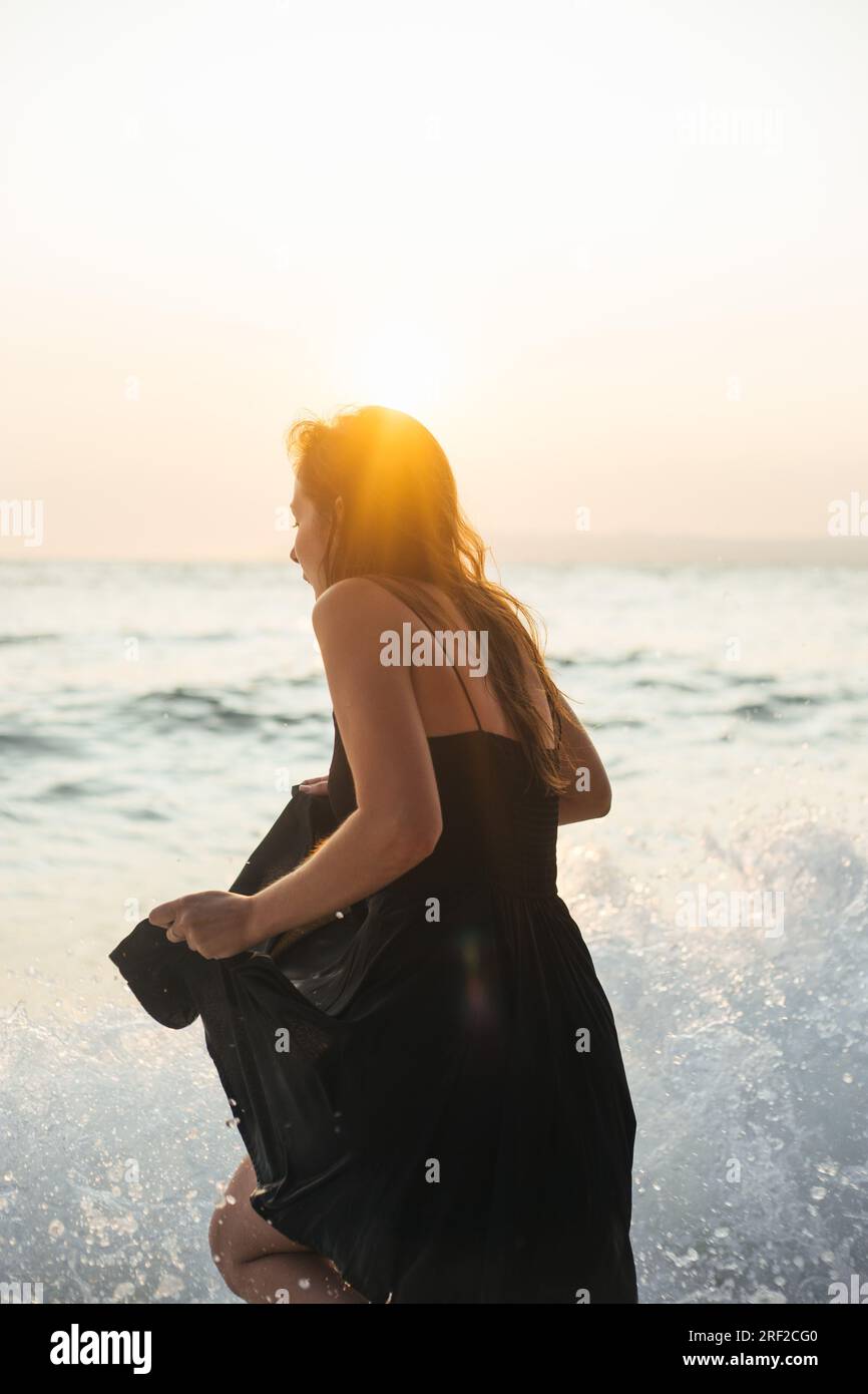 Giovane donna con vestito estivo nero che viene spruzzata dalle volte del mare Foto Stock
