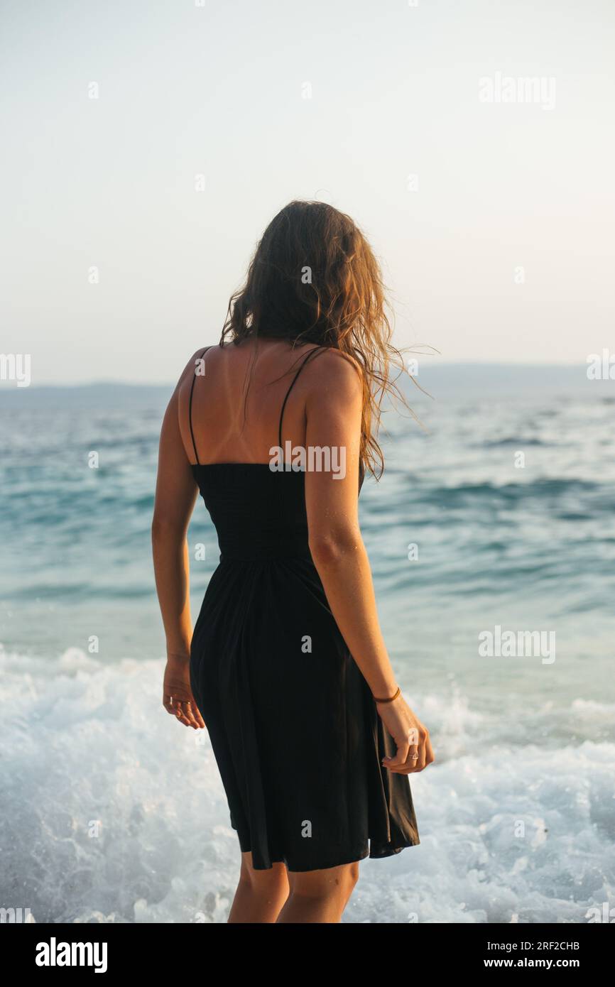 vista posteriore di una giovane donna in abito nero in piedi in fondali marini bassi Foto Stock