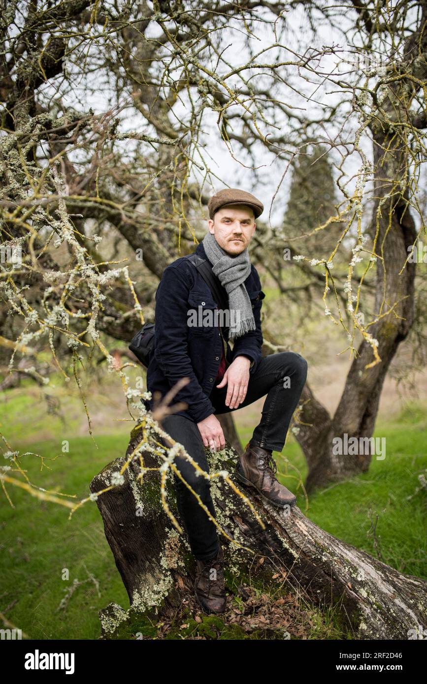 Uomo seduto su un vecchio albero caduto che guarda la macchina fotografica Foto Stock