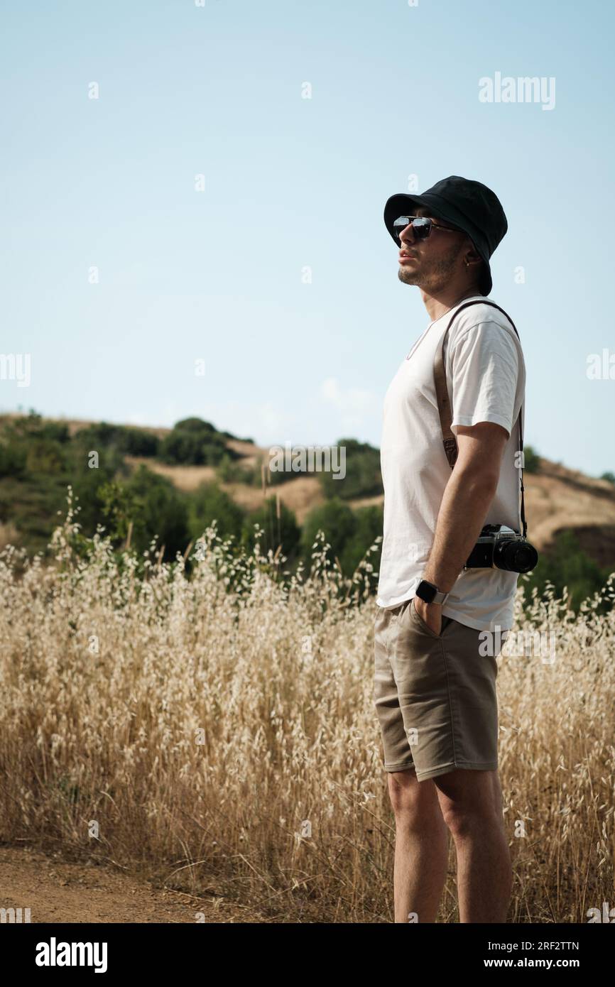 Un ritratto di un uomo di vent'anni, vestito casualmente con una t-shirt bianca, pantaloncini beige, occhiali da sole e un berretto da marinaio nero, con una classica fotocamera slun Foto Stock