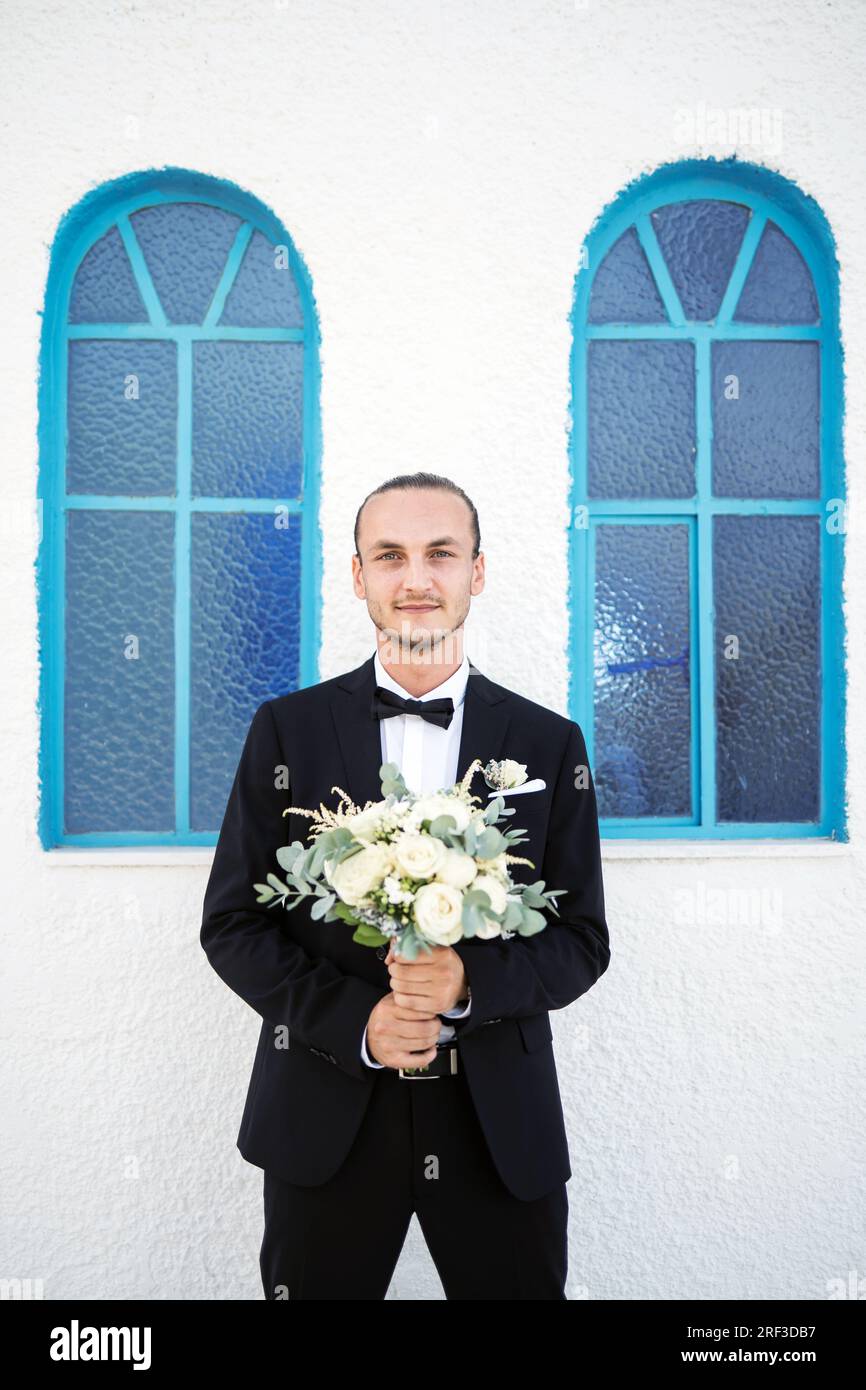 Stilvoller Bräutigam wartet vor der Kirche auf die Braut mit einem Blumenstrauß Foto Stock