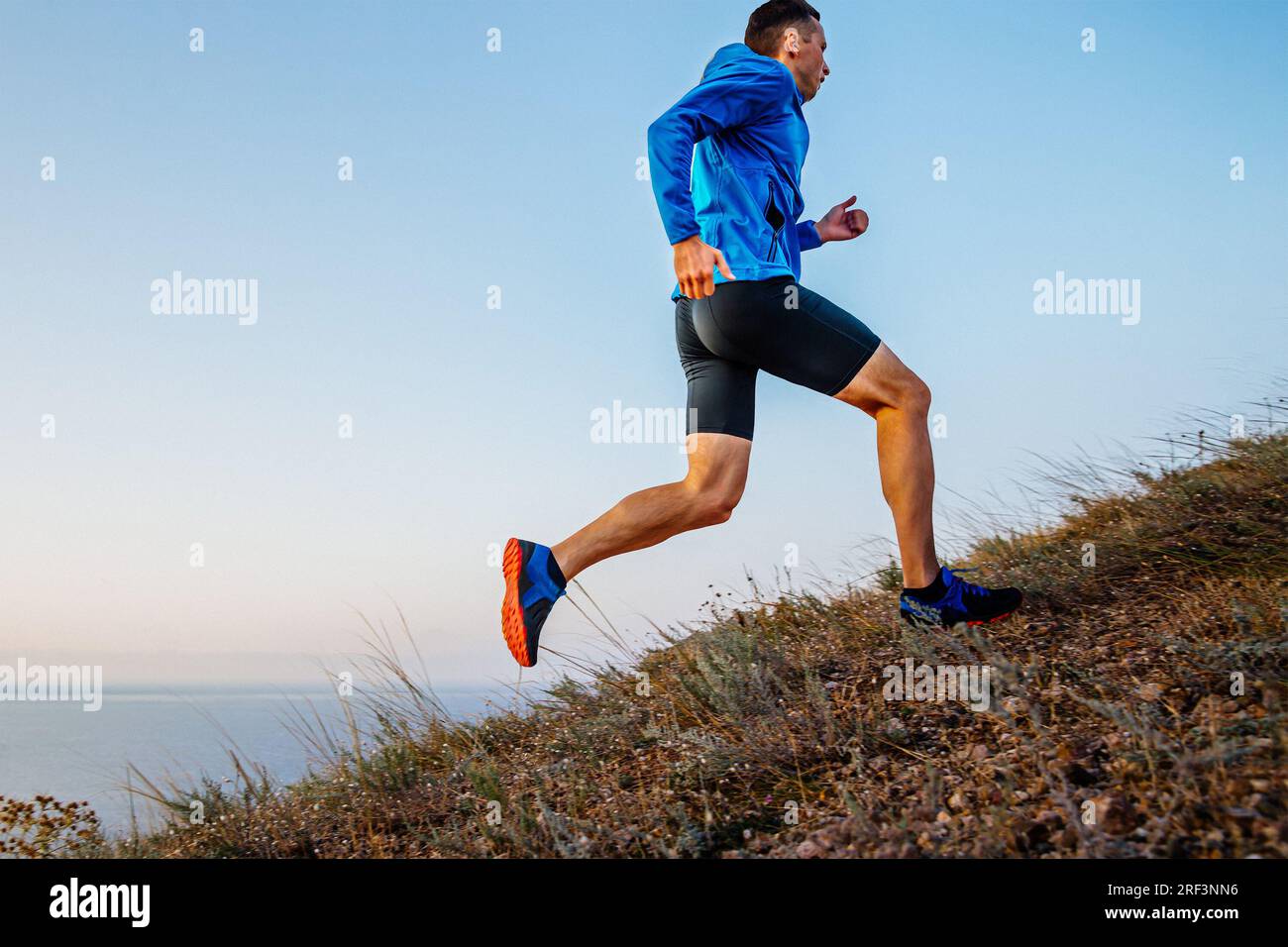 atleta maschile con vista laterale che corre in salita con giacca blu e tights neri, sfondo di cielo e mare Foto Stock