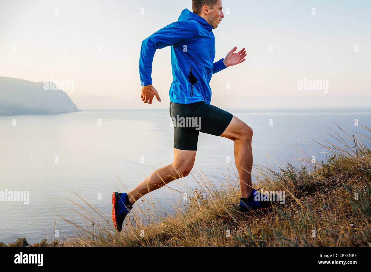 atleta atleta maschile che corre in salita con giacca blu e tights neri, sfondo di cielo e mare Foto Stock