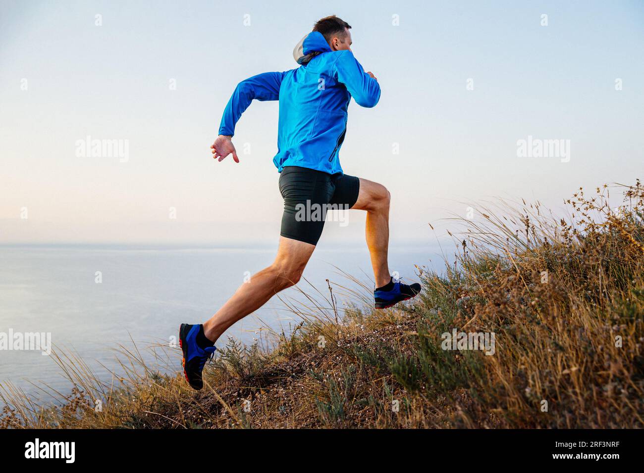 corridore maschile che corre in salita con giacca blu e collant neri, sfondo di cielo e mare, erba secca del sentiero Foto Stock