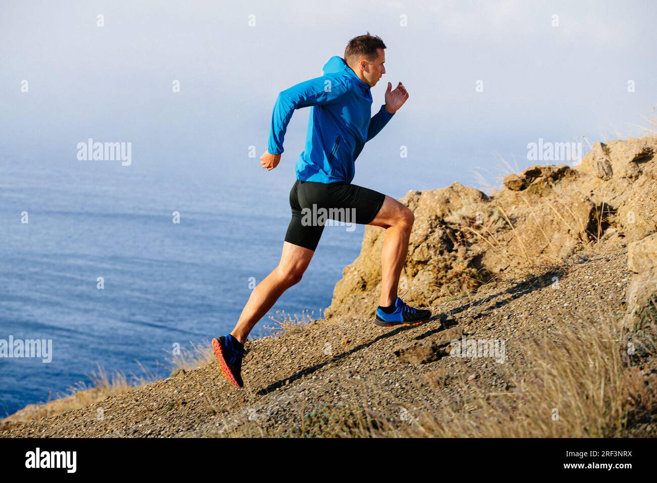 atleta runner che corre in montagna in salita con giacca blu e tights neri, sfondo di mare Foto Stock