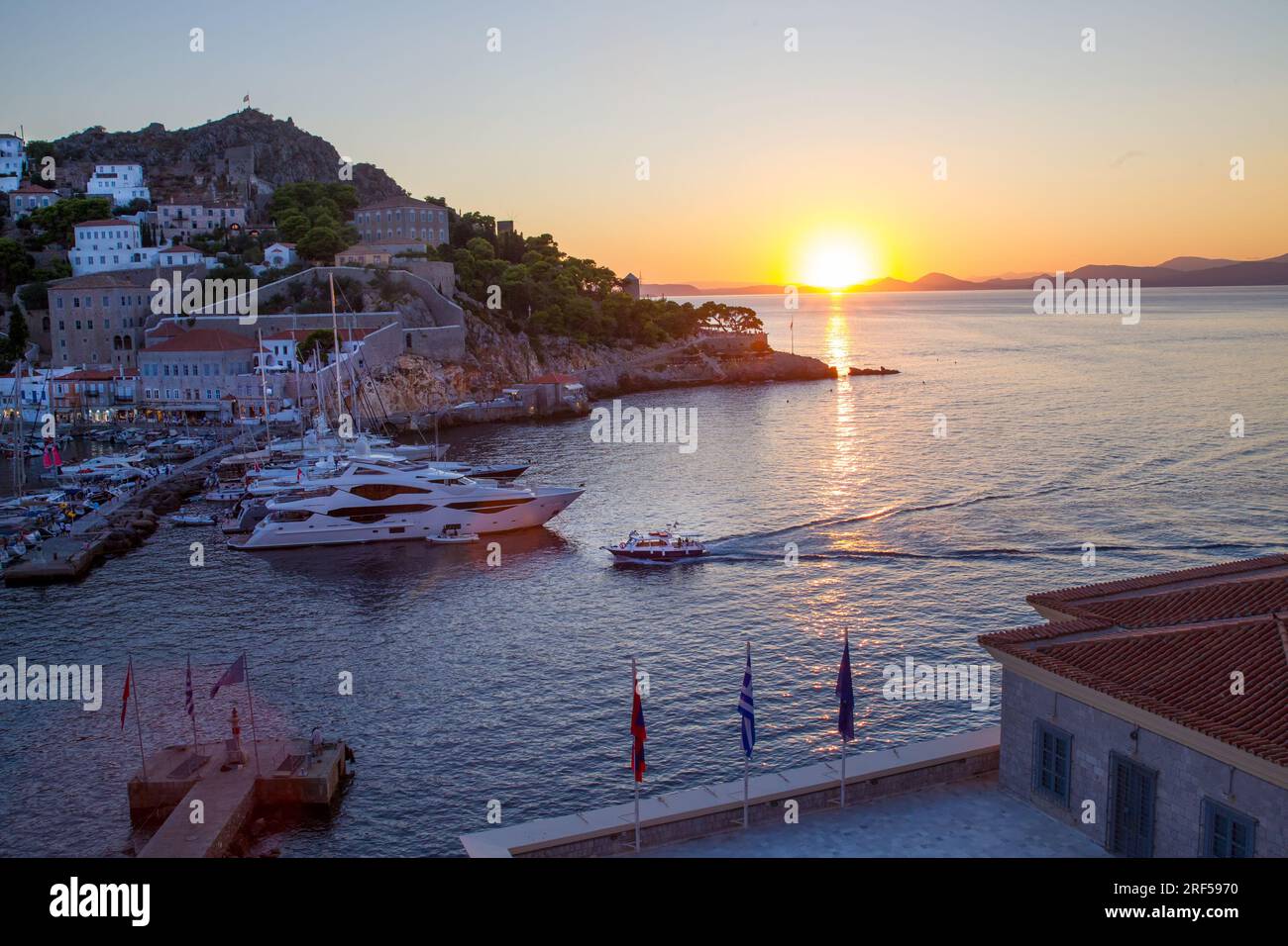 Baia di Hydra in un fantastico tramonto. Una piccola barca (taxi d'acqua) dalla Grecia continentale si avvicina al porto. Grandi yacht nei parcheggi. Vacanze idilliache Foto Stock