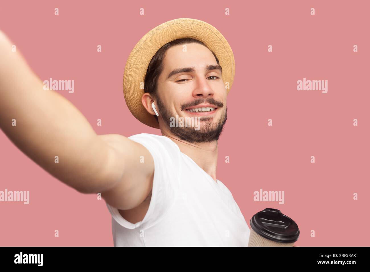 Ritratto di un uomo barbato allegro e allegro in maglietta bianca e cappello che beve caffè in piedi, sorridendo davanti alla macchina fotografica, POV. Riprese in studio per interni isolate su sfondo rosa. Foto Stock