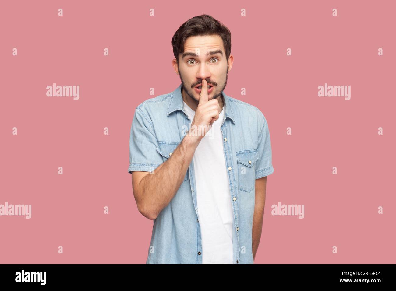 Ritratto di un uomo barbuto segreto e sbalordito in camicia blu casual in piedi fa gesto tranquillo, chiede di rimanere in silenzio, pettegolezzi su qualcosa. Riprese in studio per interni isolate su sfondo rosa. Foto Stock