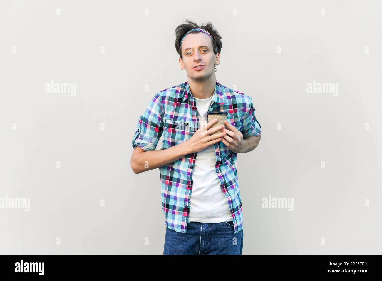 Bell'uomo sorridente che tiene il caffè da asporto davanti al petto, guarda la macchina fotografica, ama il caffè, indossa camicia a scacchi blu e fascia. Riprese in studio in interni isolate su sfondo grigio. Foto Stock