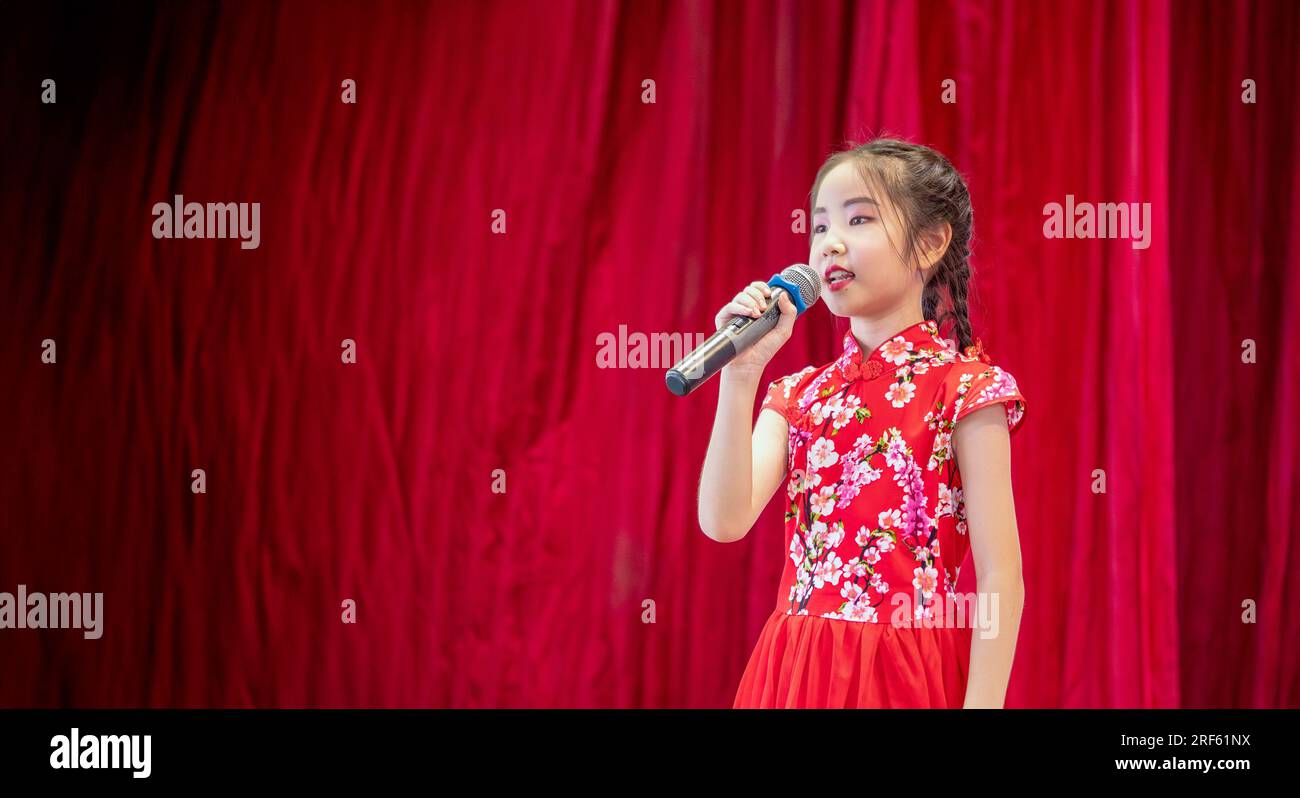 La bambina asiatica canta una canzone sul palco durante la giornata di attività scolastica, vestita in stile Qipao, sfondo rosso tenda Foto Stock