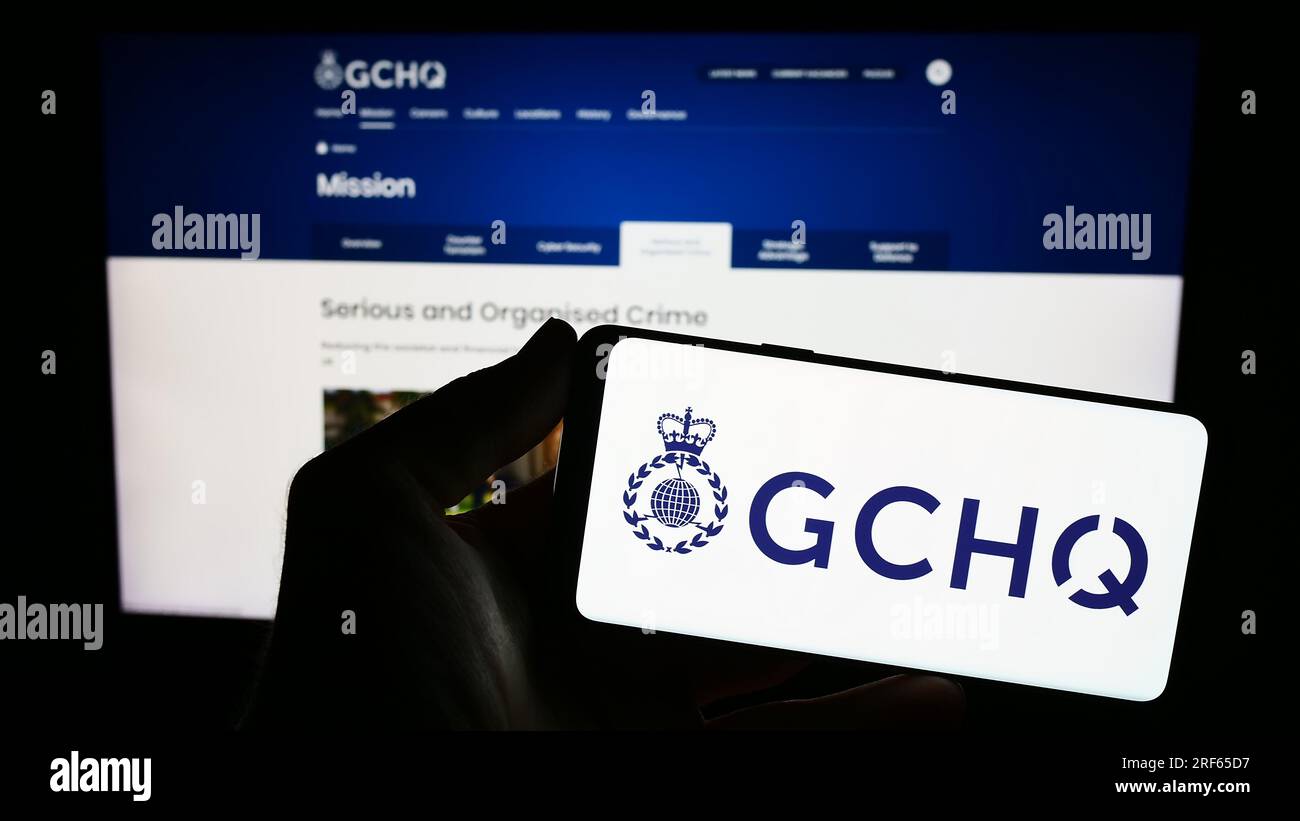 Persona che possiede il cellulare con il logo della sede centrale delle comunicazioni governative (GCHQ) sullo schermo davanti alla pagina Web. Concentrarsi sul display del telefono. Foto Stock
