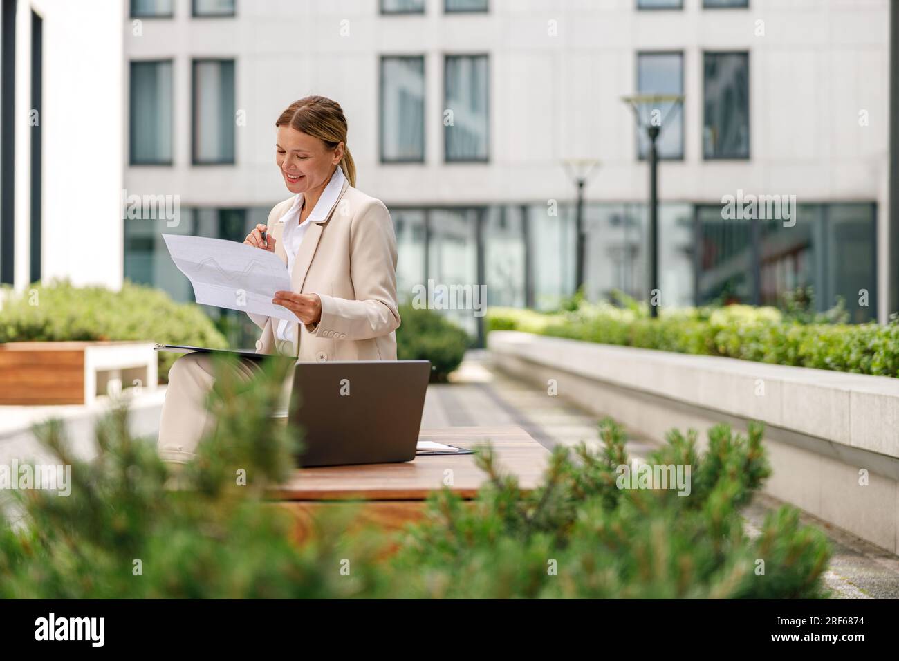 Analista donna che lavora con documenti e utilizza un notebook seduto all'esterno su un edificio moderno Foto Stock