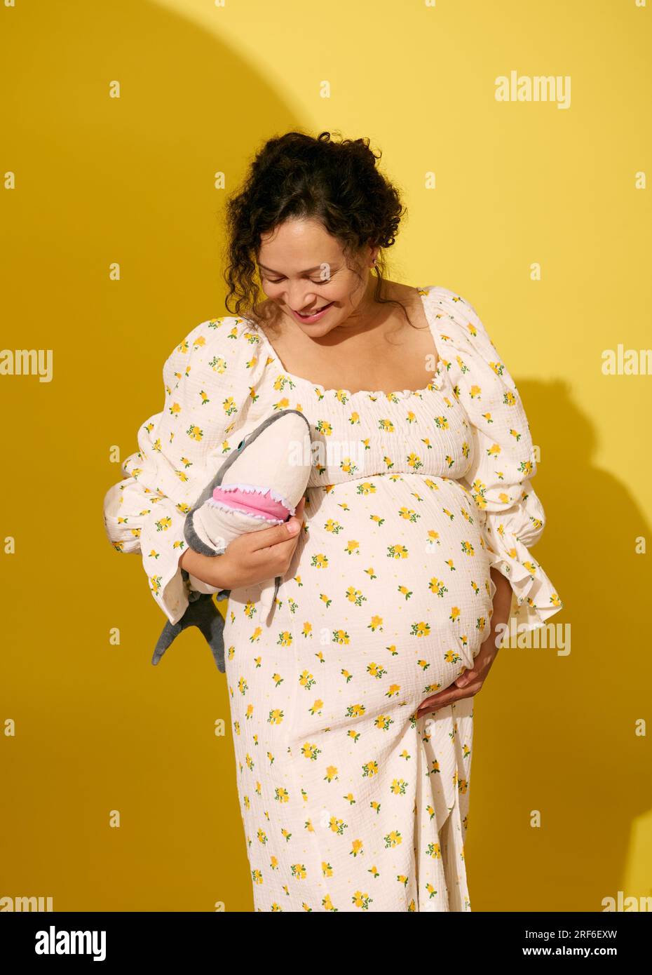 Allegra donna incinta, riccia e multietnica sorridente, con in mano un peluche giocattolo di squali, isolata su sfondo giallo dello studio. Concetto di maternità e gravidanza Foto Stock