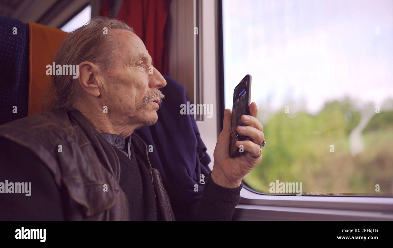 Uomo anziano che viaggia in treno e scatta foto del paesaggio attraverso la finestra utilizzando uno smartphone Foto Stock