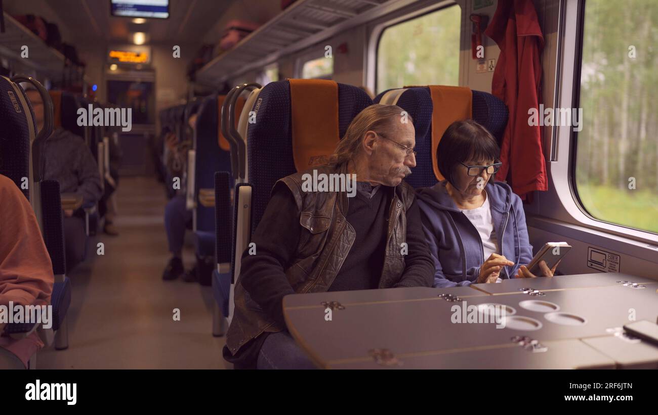 La coppia anziana viaggia in treno, la signora tiene in mano un telefono cellulare, entrambi guardano lo smartphone e parlano tra loro Foto Stock