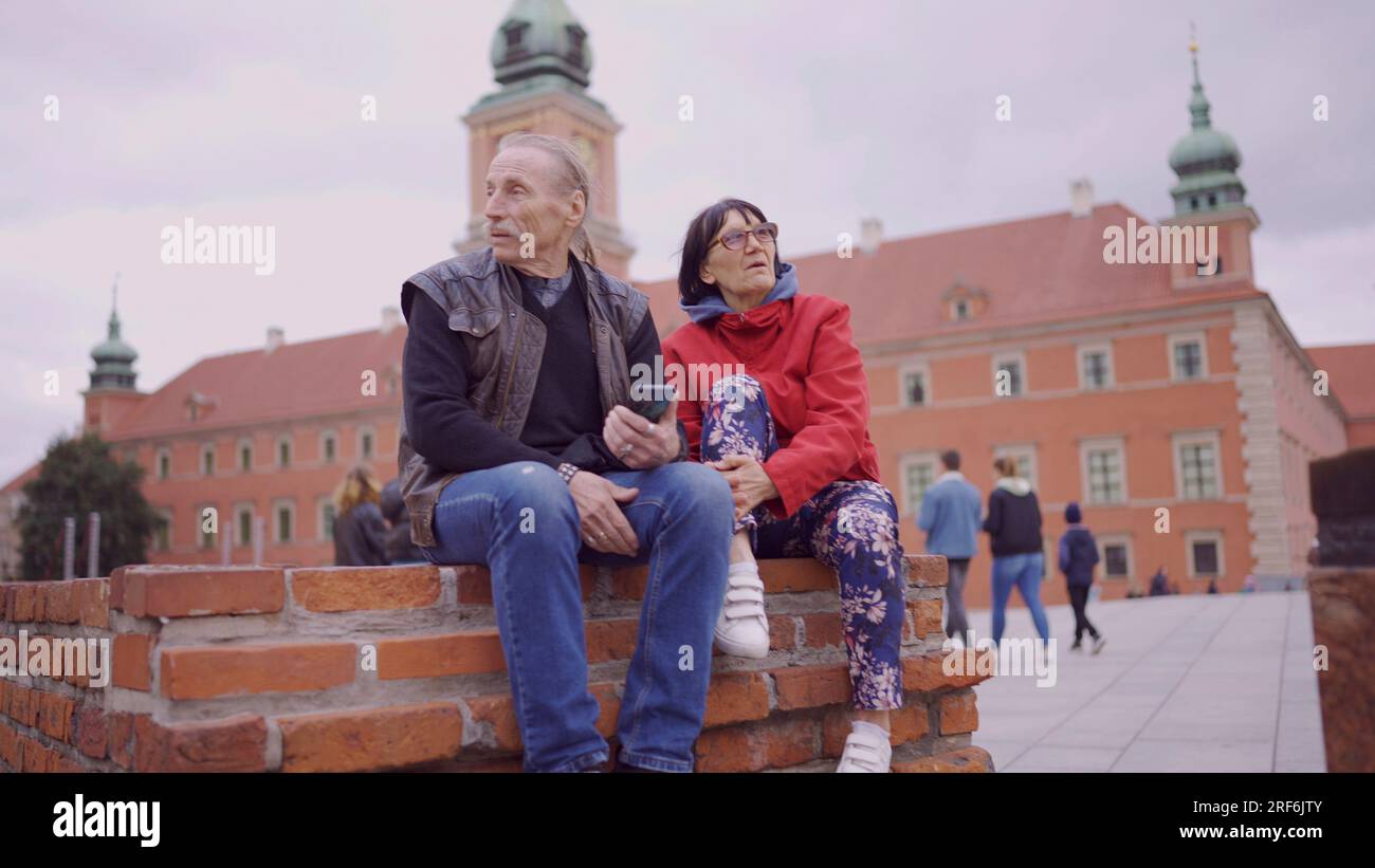 Una coppia di turisti anziani è seduta e parla nel centro storico di una vecchia città europea, senior ha uno smartphone in mano. Piazza del Palazzo, Foto Stock