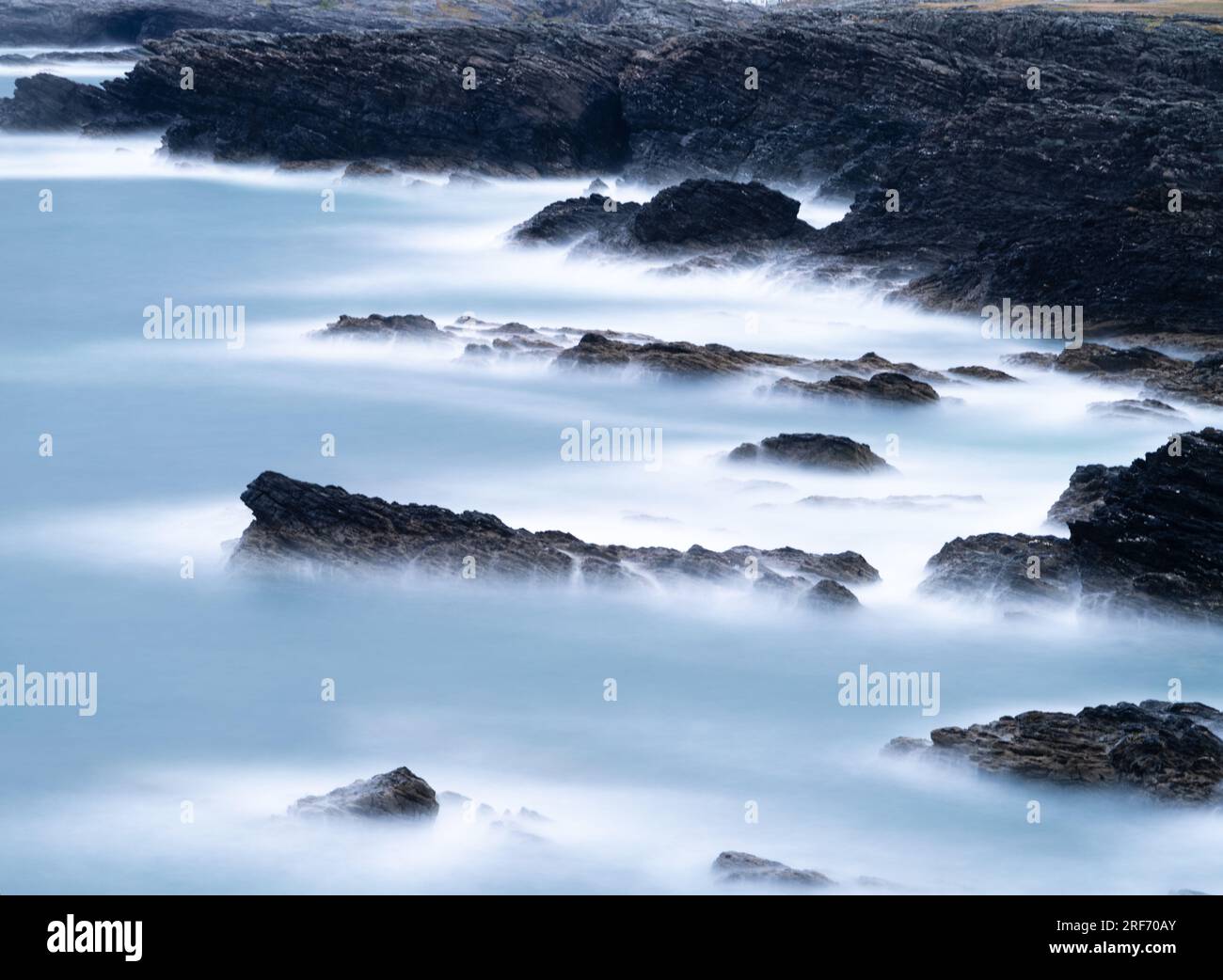 Lunga esposizione di una linea costiera rocciosa. Il mare ha un leggero colore metallico con bordi bianchi nebbiosi contro le rocce nere. Foto Stock