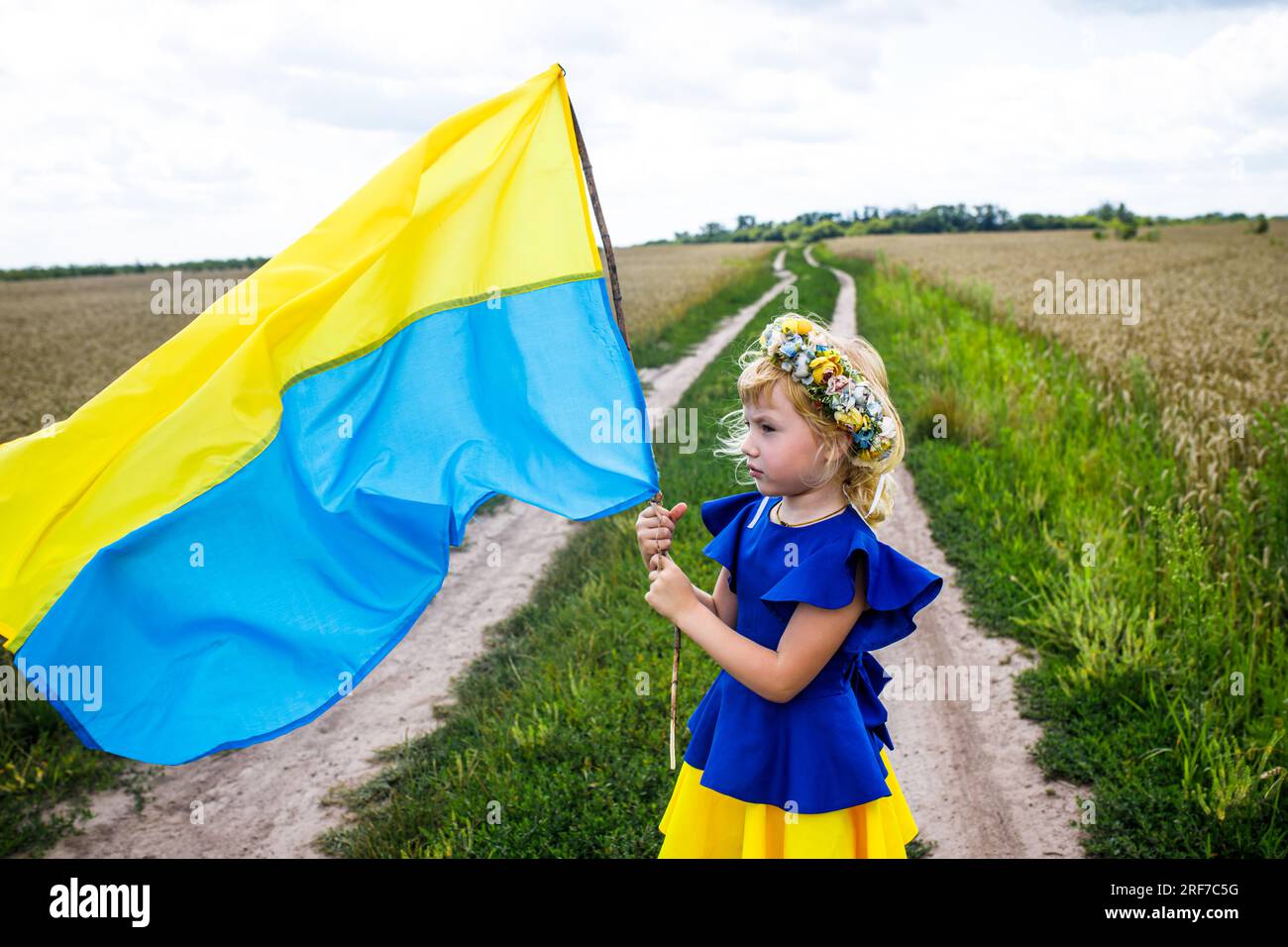 La bandiera dell'Ucraina è sulle spalle di una bambina Ucraina. Un bambino ucraino felice in Ucraina libera senza guerra corre lungo una strada rurale nel grano Foto Stock